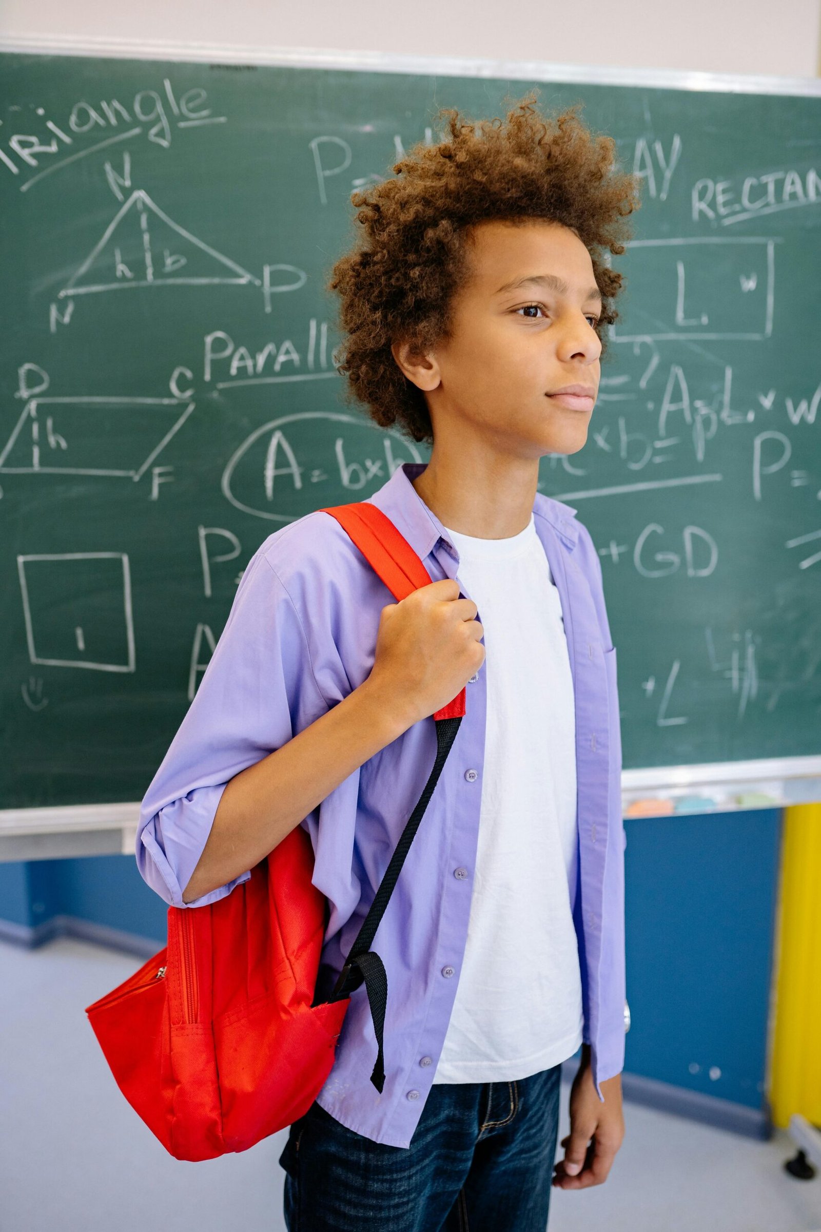 Teenage student with red backpack standing in front of a blackboard in classroom.